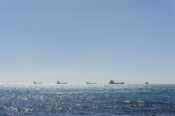 Fleet of cargo ships and vessels anchored in the open sea on the horizon line under a clear blue sky with sun glares on water © Natalia