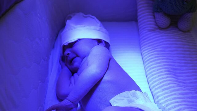 Top down view of a newborn baby sleeping in a special bassinet under a blue ultraviolet lamp, undergoing phototherapy at home for neonatal jaundice treatment to reduce bilirubin levels