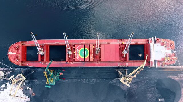 Red bulk carrier moored beside black cargo stockpile. Direct overhead view captures open holds, deck cranes and a helipad aligned along the docked vessel.