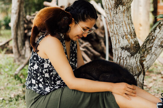 Mujer feliz al aire libre interactuando con un peque&ntilde;o mono y un coat&iacute; con cari&ntilde;o, celebrando la conexi&oacute;n con la fauna en el D&iacute;a Mundial de los Animales.