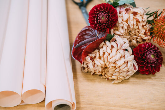Flower craft. Bouquet preparing. Red dahlias chrysanthemums color anthurium protea near wrapping paper on wooden table in professional florist shop.