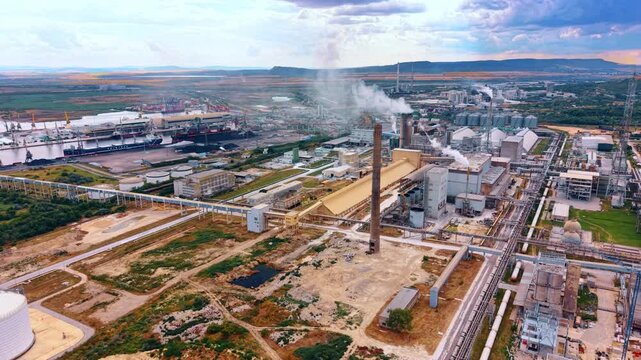 Processing plant and cargo port connected by conveyors. Elevated view includes smokestacks, pipelines, warehouses, docked ships and bulk material piles.