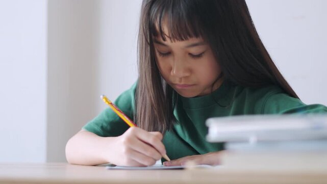 Close up selective focus of a concentrated Asian kid, a young Thai girl or Chinese student, diligently doing homework and writing with a pencil in a notebook. Education, learning, and study concept.