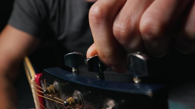 A close-up view shows a person fingers carefully adjusting a metallic tuning peg on the headstock of an acoustic guitar. The dark background highlights the intricate details of the hardware.