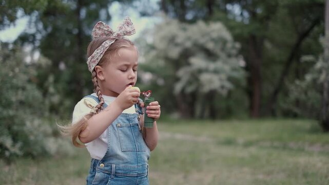 Cute little girl blowing soap bubbles in summer park with boy playing in background