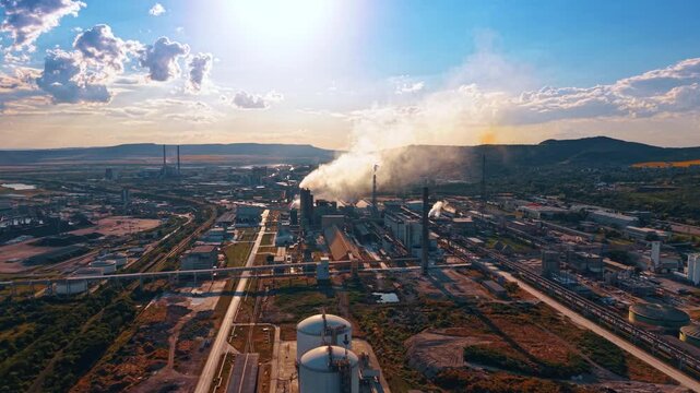 Industrial complex with steam plumes under direct sunlight. Storage tanks, pipelines, production halls and rail lines extend across a broad valley.