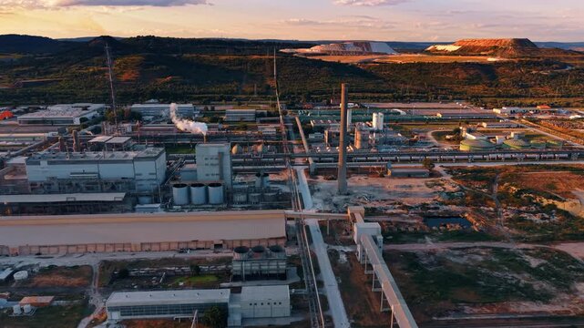 Industrial processing buildings and pipelines beneath terraced quarry. Drone view of factory halls, stacks and pipe bridges with excavated plateaus across wooded hills.