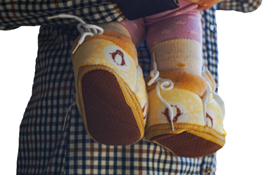 A detailed shot of a baby's small feet wearing soft yellow patterned booties with white laces. The baby is being held by an adult wearing a checkered shirt, isolated on a clean white background. Perfe