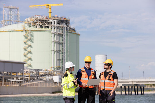 Team of engineers wearing safety equipment working together on a construction site with a large industrial facility visible in the background