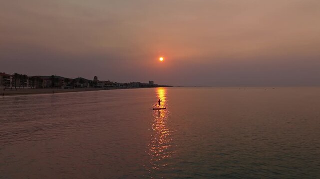 Cinematic wide shot of a person paddling on a stand-up paddleboard across a calm sea, passing directly through a bright golden sun glint on the water surface during a peaceful coastal sunset.