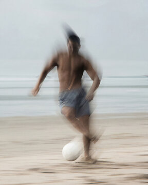 Abstract beach soccer player in motion blur: shirtless athletic man kicking white ball on sand with ocean horizon background.