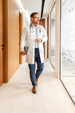 Professional doctor holding a dark tablet and wearing a white lab coat with a stethoscope around shoulders. Walking confidently through a hospital corridor