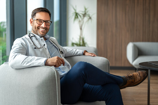 Male doctor in medical uniform and eyeglasses smiling, embodying confidence and professionalism while relaxing in a comfortable chair in a modern clinic reception area