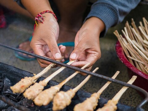 Close up of vendor hands grilling traditional Balinese sate lilit skewers over a charcoal grill at a local food stall to create an authentic Indonesian culinary