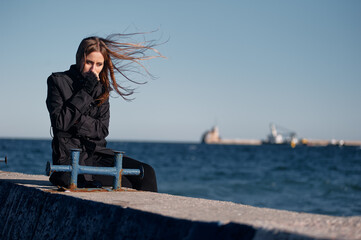 Fototapeta premium Pensive woman in a black coat sits alone on a concrete pier as wind blows her hair, watching the choppy sea and distant ships. Moody coastal portrait conveying solitude and contemplation.