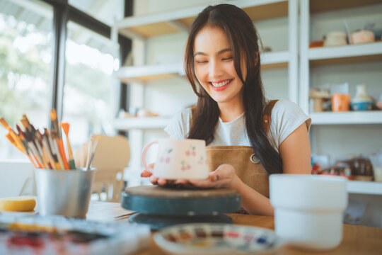 Smiling Asian woman hands showing cute handmade ceramic mug with cherry pattern at pottery workshop, female artisan crafting clay cup for creative hobby, small business entrepreneur.