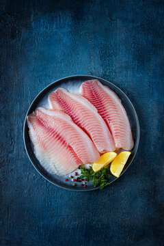 Fresh Tilapia fillet, on a plate, with lemon and herbs, blue background, no people