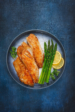 Fried fish fillets with asparagus and lemon served on a dark plate with a blue background