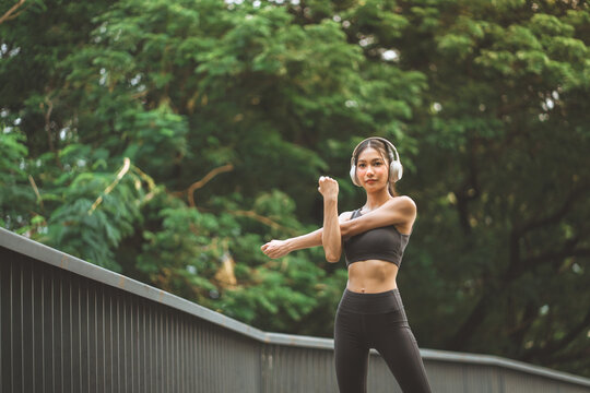 Asian woman wearing headphones and stretching arms during outdoor workout in green park, smiling female athlete listening to music and warming up before running for fitness, healthy wellness, copy spa