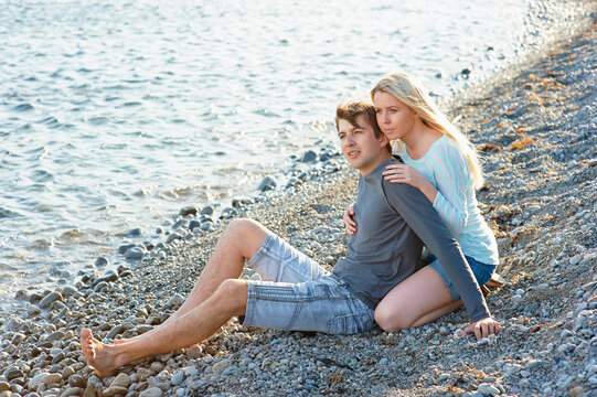 A young couple sits on a pebble beach by the sea, the woman embracing the man as they gaze toward the horizon, sharing a quiet romantic moment and relaxed summer atmosphere.