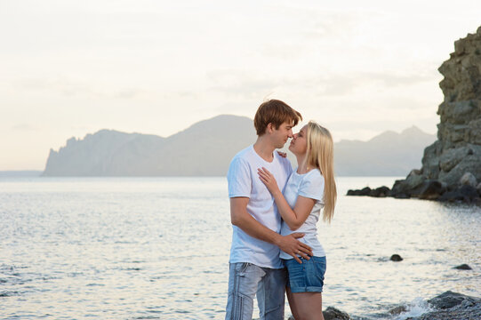 Young couple embraces and shares a kiss on a rocky seaside at sunset, enjoying a romantic moment by the water with distant mountains and calm waves, conveying love, intimacy and summer travel.