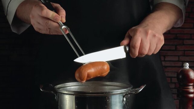 Chef in black apron using knife and fork to drop sausages into hot pan, demonstrating cooking process in kitchen with rustic brick wall and kitchen utensils in background