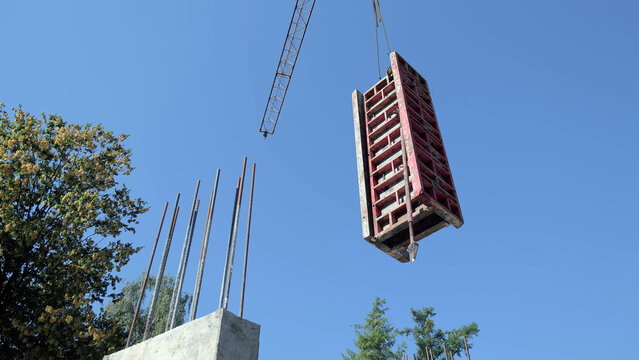 Crane Lifting Concrete Formwork at Construction Site. Crane lifting red concrete formwork above exposed rebar at a construction site under a clear blue sky.