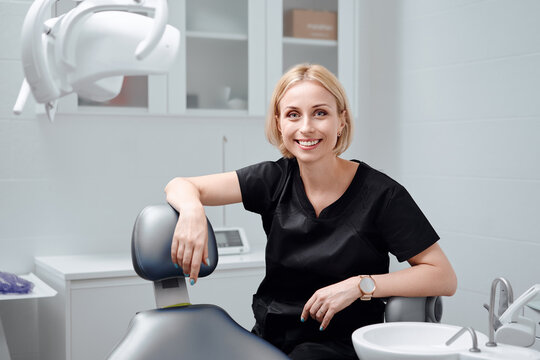 Young blonde woman dentist posing at modern dental cabinet next to dental chair cheerfully smiling at camera. Pretty stomatologist at dental clinic