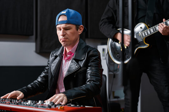 Musician seated at a keyboard playing seriously, accompanied by a guitarist in a modern studio environment. The scene portrays dedication to music and a professional creative process.