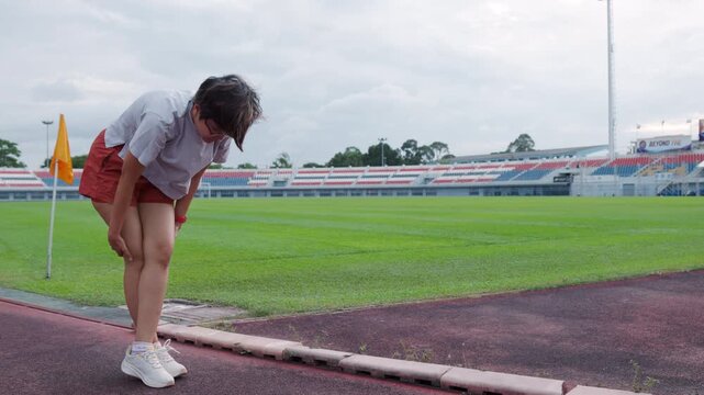 Authentic shot of an active Asian woman stretching on a running track beside a green soccer field. She bends down, providing excellent copy space for your health and fitness project backgrounds.