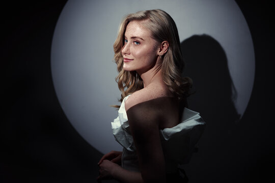 Elegant blonde woman posed in a moody studio portrait with dramatic lighting, soft smile and visible freckles. The image conveys confidence, glamour, intimacy and cinematic fashion