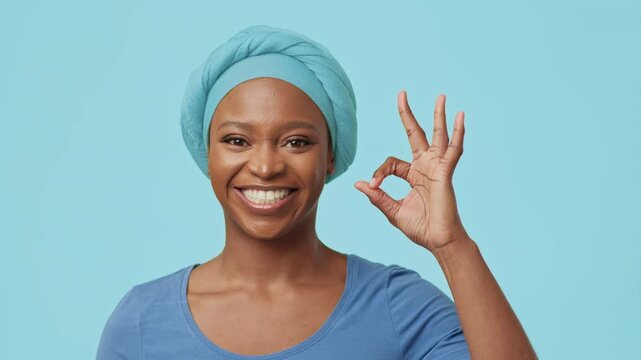 Surprised African American Woman In Blue Top Expressing Emotion With Open Mouth And Hand Gesture Of Okay Sign, Standing Against Light Blue Background, In Studio Setting. Happiness Concept.