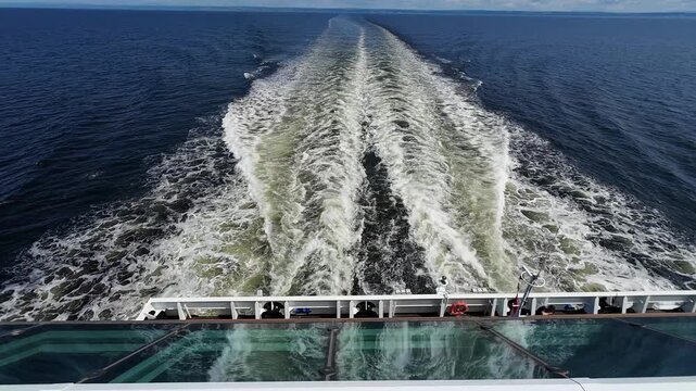 Rear view of a cruise ship sailing across open water with twin ocean wakes reflected in the large window beneath the aft deck.