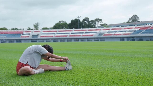 Authentic shot of an active Asian woman sitting and stretching her legs on a green soccer field. The empty stadium with colorful seats provides excellent copy space for sports backgrounds.
