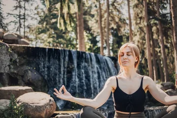 Fotobehang Lotusbloem Woman meditates in lotus position on rock in front of waterfall. Anxiety management practice, mindful living lifestyle, screen free wellness break.  © irissca