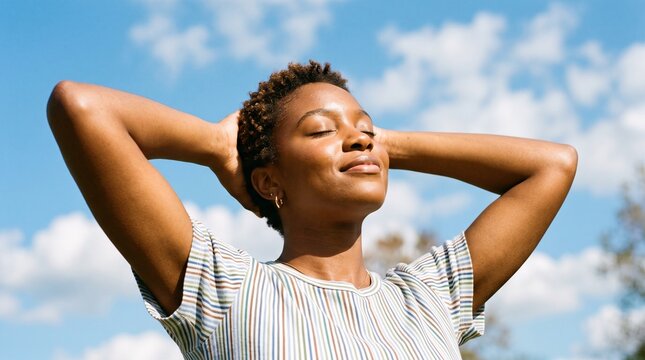 Serene young Black woman relaxing outdoors with eyes closed, enjoying sunshine and fresh air against a blue sky.