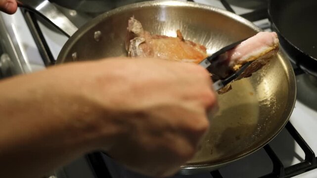 Cooking pork chops in a metallic frying pan on a kitchen stove with a wooden cutting board nearby, neutral mood, close-up side view, food preparation, cooking tutorial, brown, silver, black.