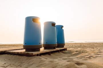 Naklejka premium Three blue trash bins sit on a wooden platform on the beach. The sun sets in the background, bathing the sand in a warm glow. The concept is about cleaning, ecology, and recycling.