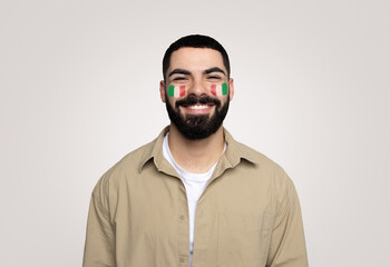 Fototapeta premium Happy Young Male, Middle Eastern, Celebrating National Pride With Italian Flag Face Paint. Posing Cheerfully In Studio Against Plain Background, Expressing Joy And Enthusiasm For Culture.
