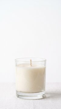 A simple white candle in a clear glass jar with a natural fiber wick centered on a textured white surface