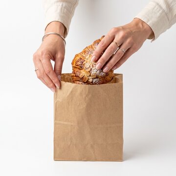 Woman's Hands Placing Almond Croissant Into Brown Paper Bag With Powdered Sugar Dusting