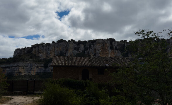 serene rustic stone house in rugged landscape with rocky cliffs and cloudy sky, ground-level perspective looking up at the house and cliffs, rural architectural photography destination.