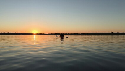 Obraz premium Kayaker Moves on Water as Sun Sets Over the Horizon Near Wooded Area