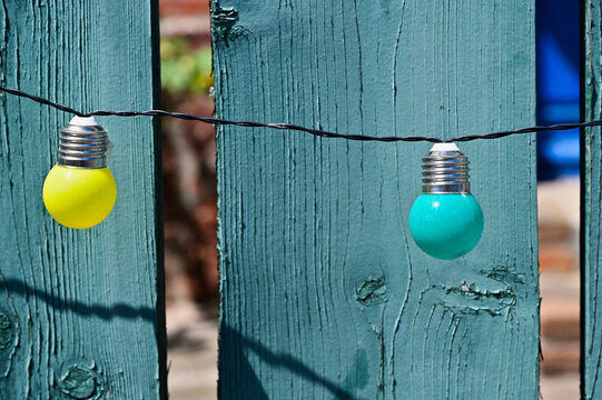 decorative small colorful light bulbs suspended on electrical wire in front of old painted blue wooden fence planks or pickets. abstract view. soft grassy background seen through gaps. home concept