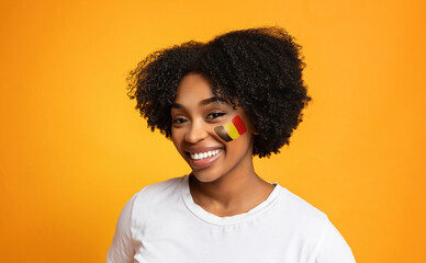 Fototapeta premium Happy Young Black Woman Smiling With Flag Painted On Face, Standing Against Yellow Background. Excited About Cultural Celebration In Studio. Represents Joy And Pride In Identity, Lifestyle Concept.
