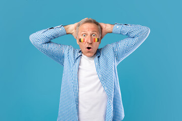 Fototapeta premium Surprised Elderly Caucasian Male With Face Paint Showing Excitement With Hands On His Head, Standing In Front Of Blue Studio Background, Celebrating Sports Event Or National Holiday.