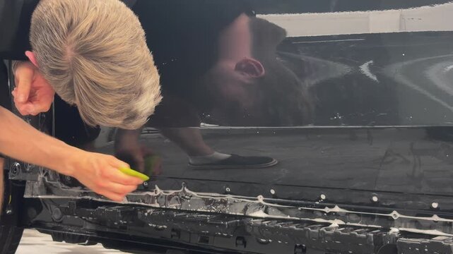 Professional car wrap installer applying a glossy transparent polyurethane paint protection film PPF on a vehicle body panel inside a premium auto detailing studio workshop.
