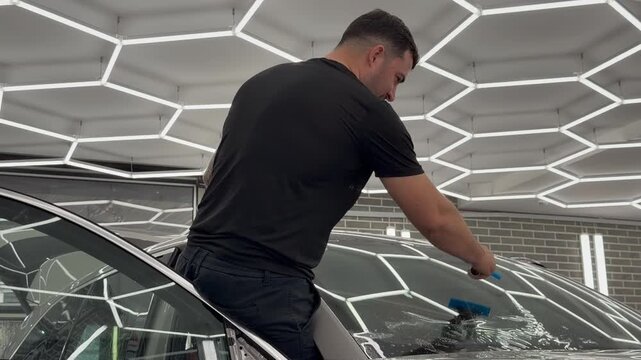 Professional car wrap installer applying a protective window film on a vehicle windshield glass to protect it from stone chips, scratches, and mechanical impacts.

