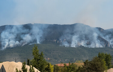 Fototapeta premium Distant view of wildfire spreading through forested mountain landscape.