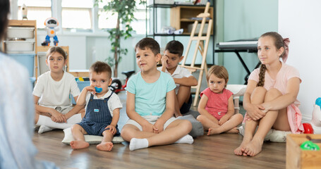 Fototapeta premium Happy children of different ages sitting on floor and listening to female teacher in playroom